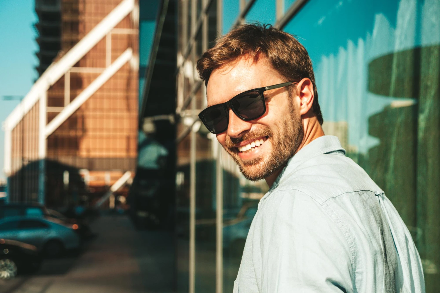 Portrait handsome smiling stylish hipster man dressed blue shirt fashion male posing street background near skyscrapers sunglasses